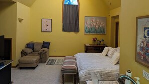 Un dormitorio con techo inclinado, una ventana con cortina, una cama, una silla, un escritorio y un televisor.