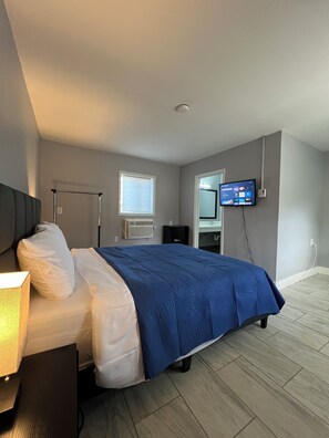 A modern bedroom with a bed, bedside lamp, air conditioning unit, and a mounted TV displaying a streaming service interface. - Nest Inn (Watertown)
