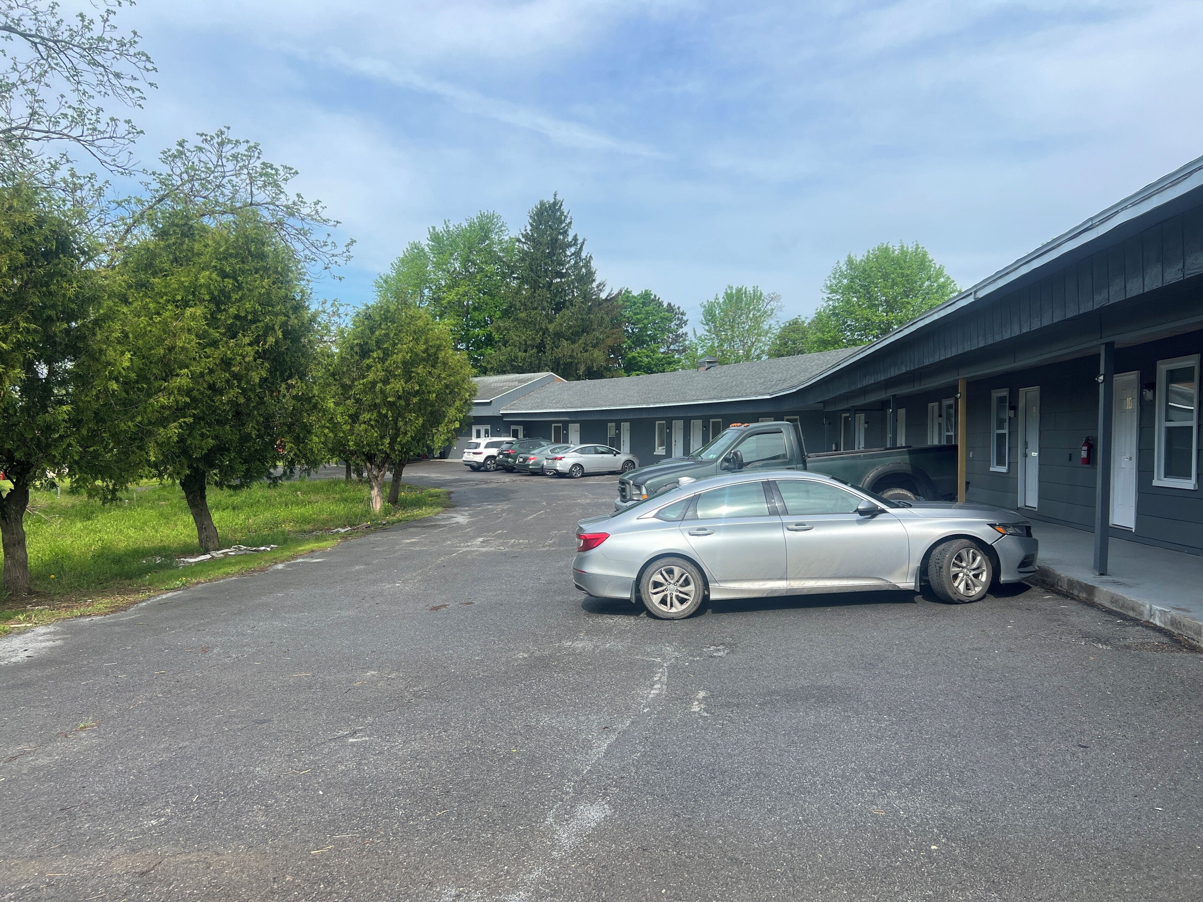 A motel with a parking lot, several cars parked, and trees lining the street.