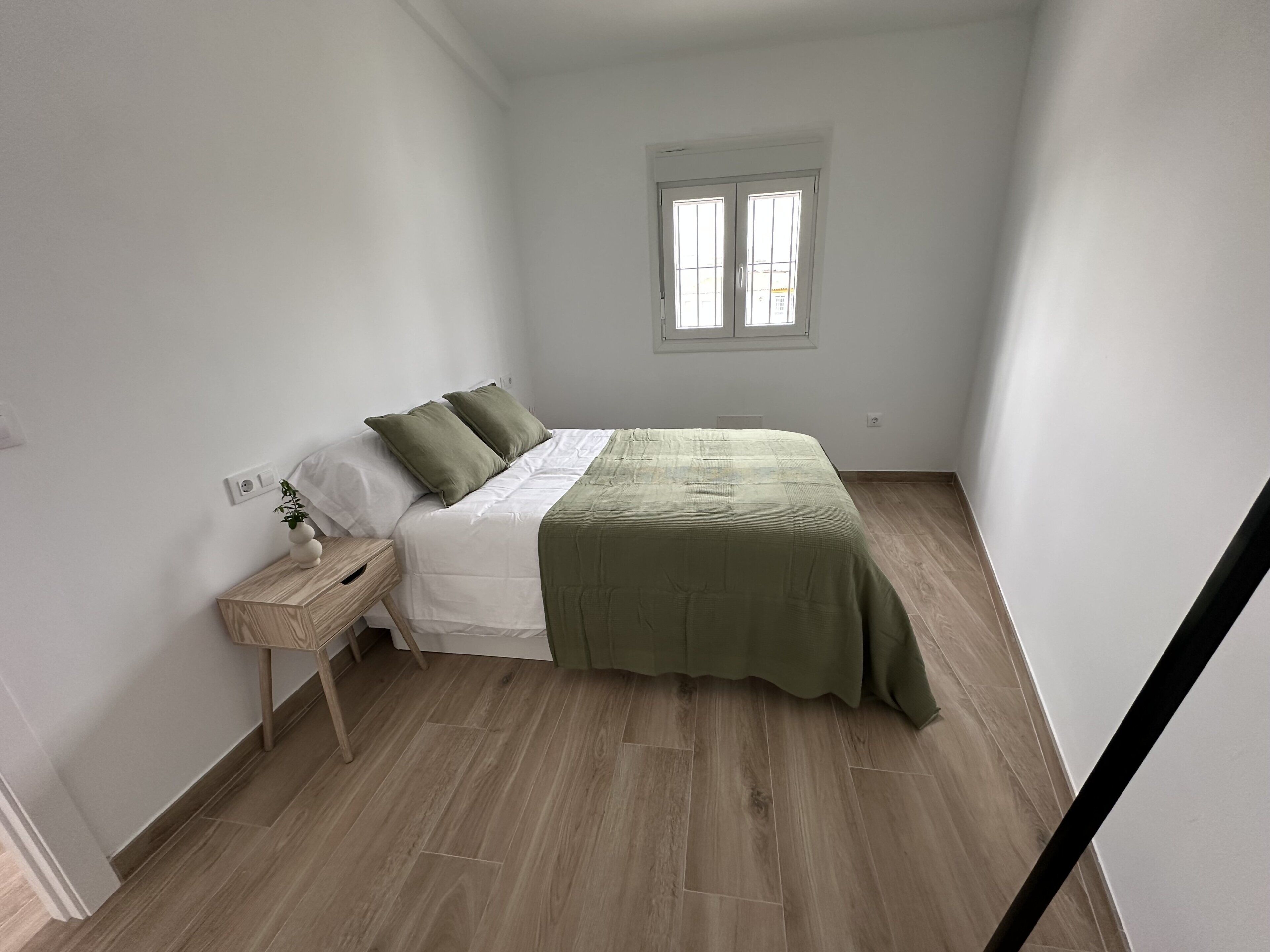 Un dormitorio con cama, mesita de noche, ventana y piso de madera.