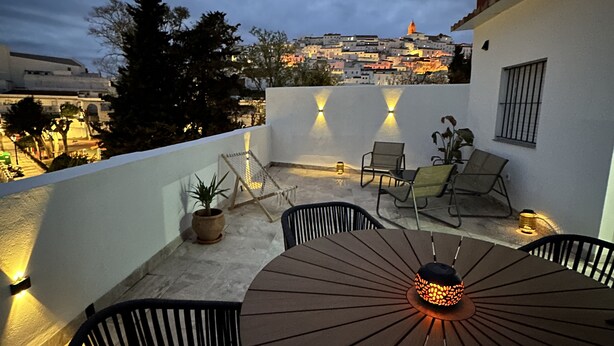 A rooftop terrace with a table and chairs, a potted plant, and a view of a cityscape at dusk. - Private Terrace, Chillout & Outdoor Dining (Alcala De Los Gazules)