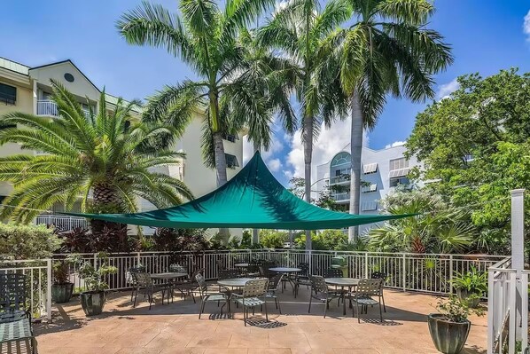 An outdoor patio area with tables and chairs, surrounded by palm trees and potted plants.