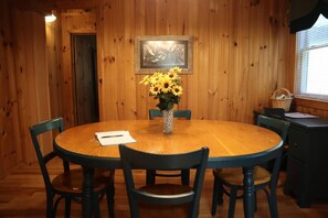 A wooden-paneled room with a round table, six chairs, a vase with sunflowers, a framed picture on the wall, and a window with blinds.