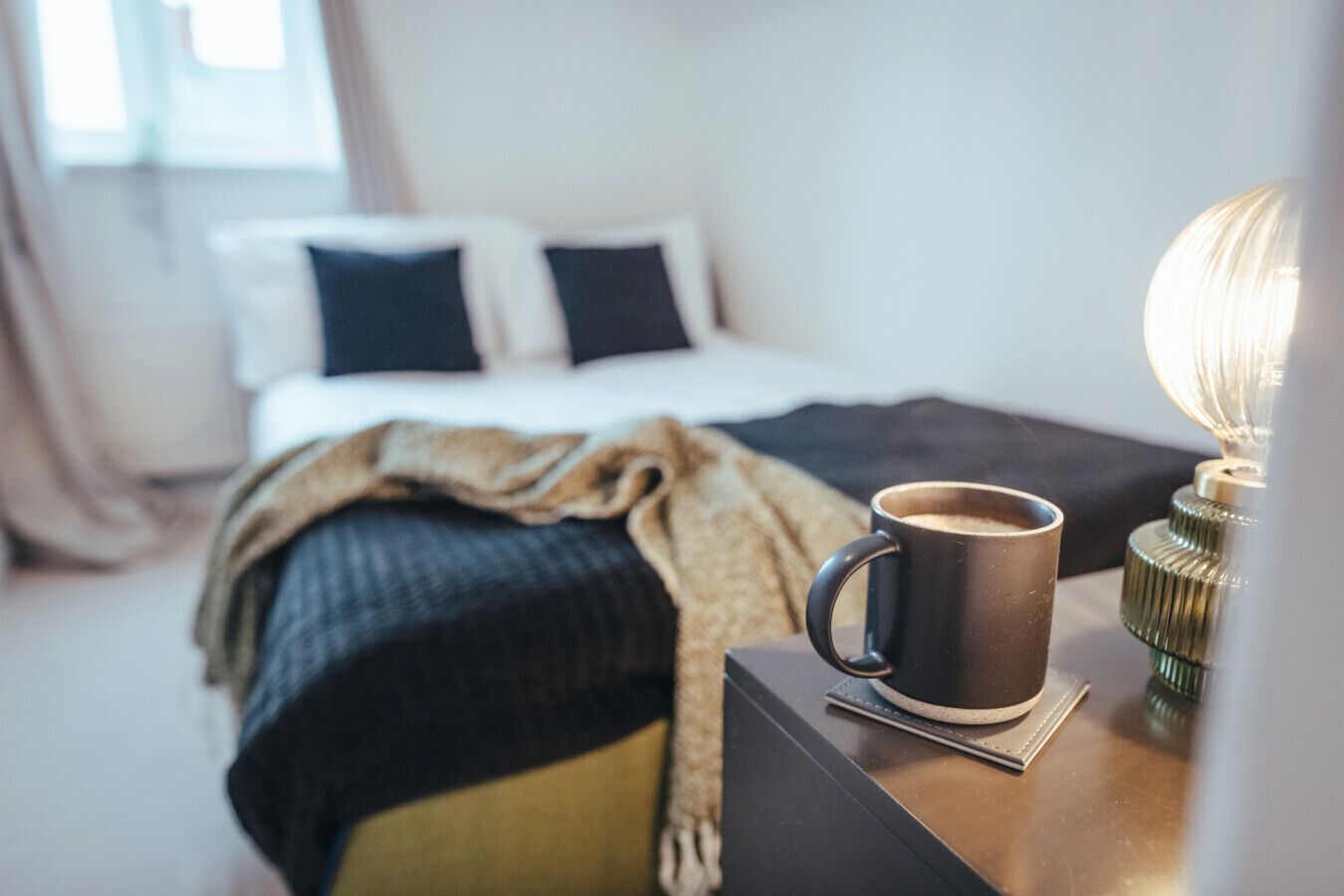 A neatly made bed with a dark blanket and pillows, a coffee cup on a side table, and a lamp with a white shade.