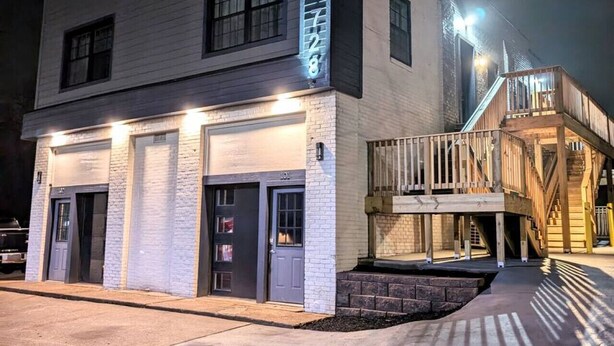 A two-story building with a garage entrance, a wooden deck, and a brick pathway leading to the entrance. - New Modern 2 BR in Excellent Location (201) (Maryville)