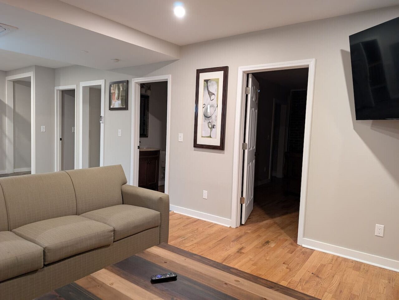 A living room with a beige sofa, a coffee table, and a flat-screen TV mounted on the wall. There are wooden floors and a door leading to another room.