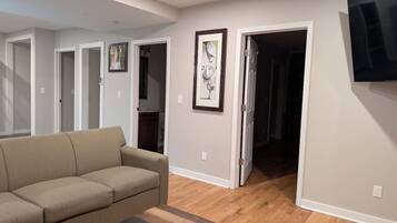 A living room with a beige sofa, a coffee table, and a flat-screen TV mounted on the wall. There are wooden floors and a door leading to another room.