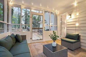 Quarto aconchegante com sofá de canto, uma mesa pequena e uma cadeira. Há uma porta de vidro deslizante que dá para um deck externo com mesa e cadeiras.