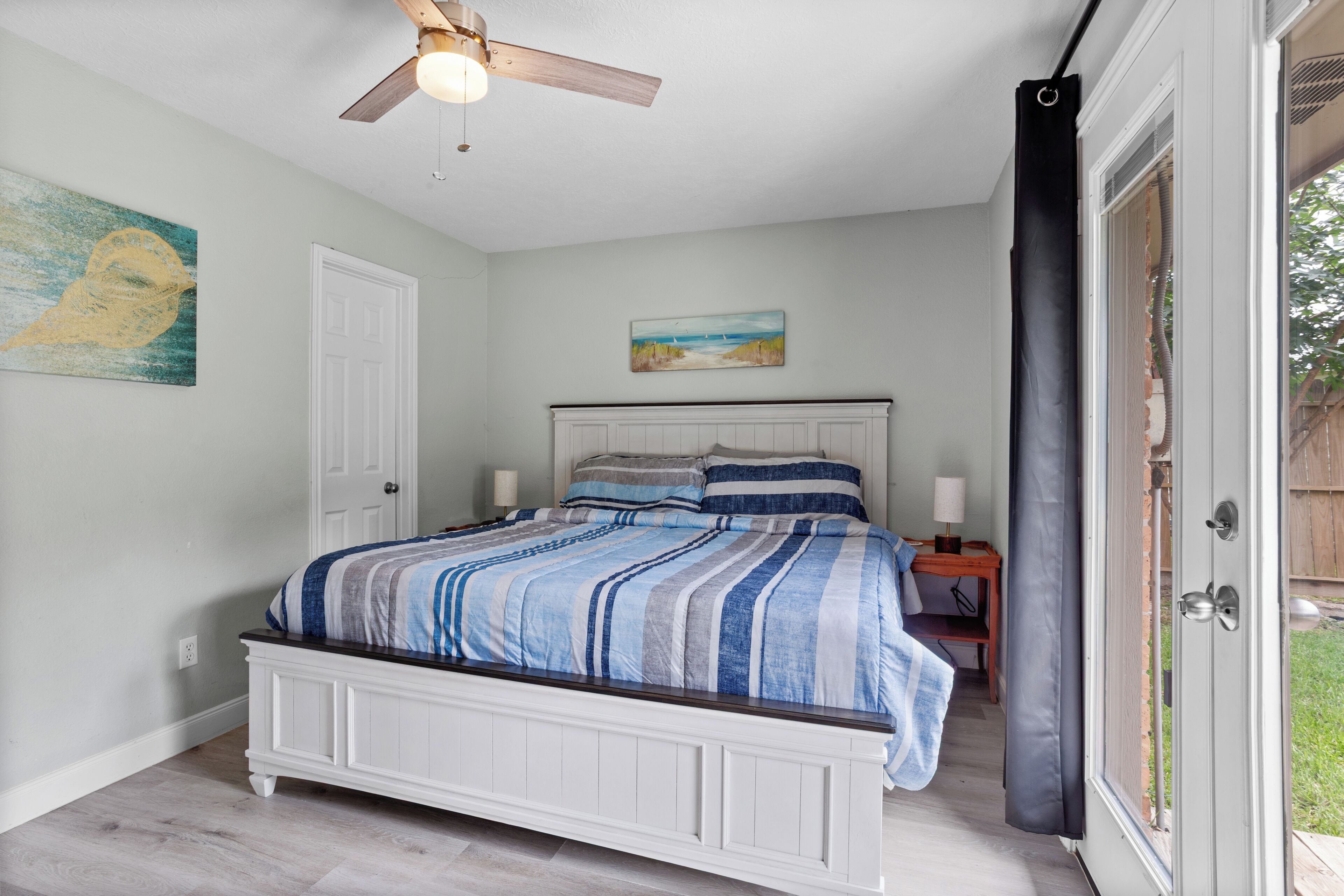 Une chambre à coucher comprenant un lit, un ventilateur de plafond, un tableau et une porte donnant sur un espace extérieur.