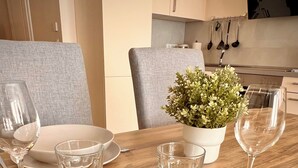 A dining table set with white plates, glasses, and cutlery, a potted plant, and a kitchen in the background. - Kleine Auszeit - Scharbeutz **incl. Parking | WiFi | Terrace with beach chair** (Scharbeutz)
