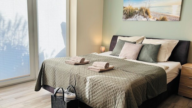 A bedroom with a bed, pillows, a bedside table, a lamp, a window, and a painting on the wall. - Kleine Auszeit - Scharbeutz **incl. Parking | WiFi | Terrace with beach chair** (Scharbeutz)