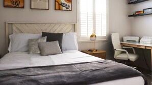 Une chambre bien rangée, avec un lit, une table de chevet, une lampe, une chaise et un bureau équipé d’une imprimante.