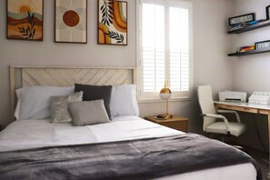 A neatly arranged bedroom with a bed, bedside table, desk, chair, and artwork on the wall.