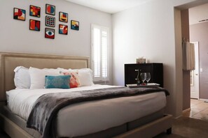 A bedroom with a bed, pillows, a bedside table with a bottle and glasses, and abstract art on the wall.