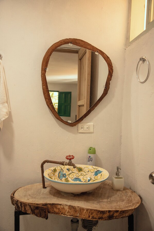 Un baño con tocador de madera, un lavabo, un espejo con marco rústico, una toalla colgada en la pared y una ventana verde visible en el reflejo.