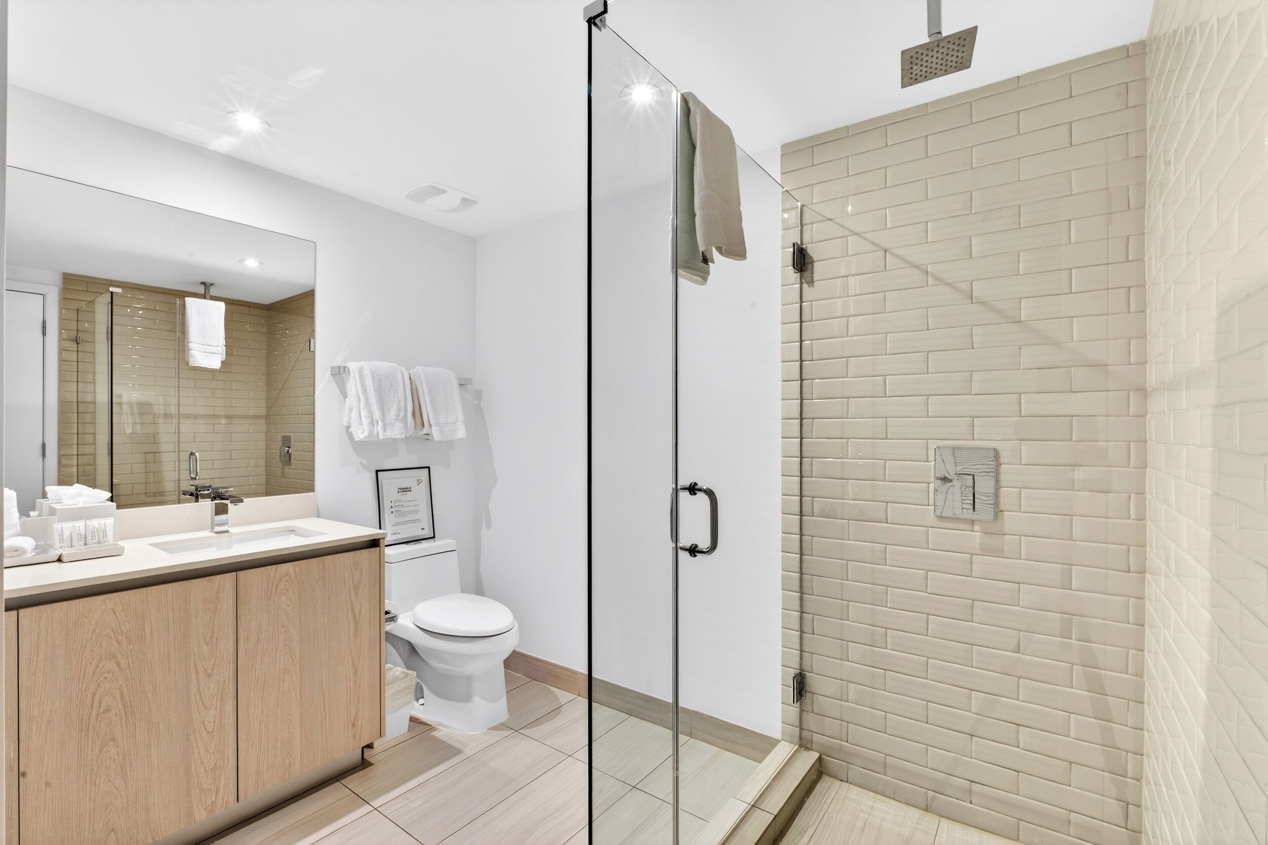 Un baño moderno con ducha de vidrio, tocador de madera, inodoro blanco y un espejo grande.