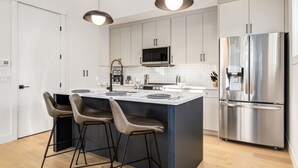 Cocina moderna con una isla central, taburetes de bar, electrodomésticos de acero inoxidable e iluminación colgante.