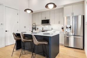 Une cuisine moderne dotée d’un îlot central, de tabourets de bar, d’appareils électroménagers en acier inoxydable et de luminaires suspendus.