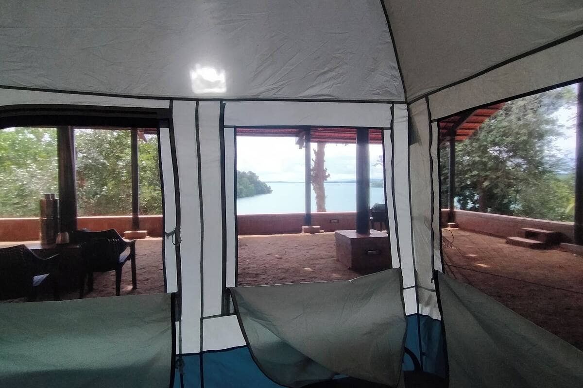 Un espace aménagé avec une tente, offrant une vue sur un lac et des arbres. Des chaises et une table sont disposées à l’extérieur.
