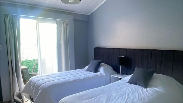 A hotel room with two beds, a large window with curtains, a ceiling light, and a view of greenery outside. - Gugasuites (Ezeiza)