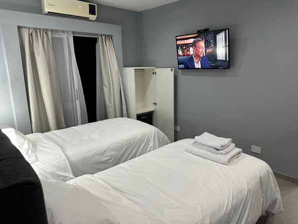 A hotel room with two beds, a television mounted on the wall, and a window with curtains. - Gugasuites (Ezeiza)