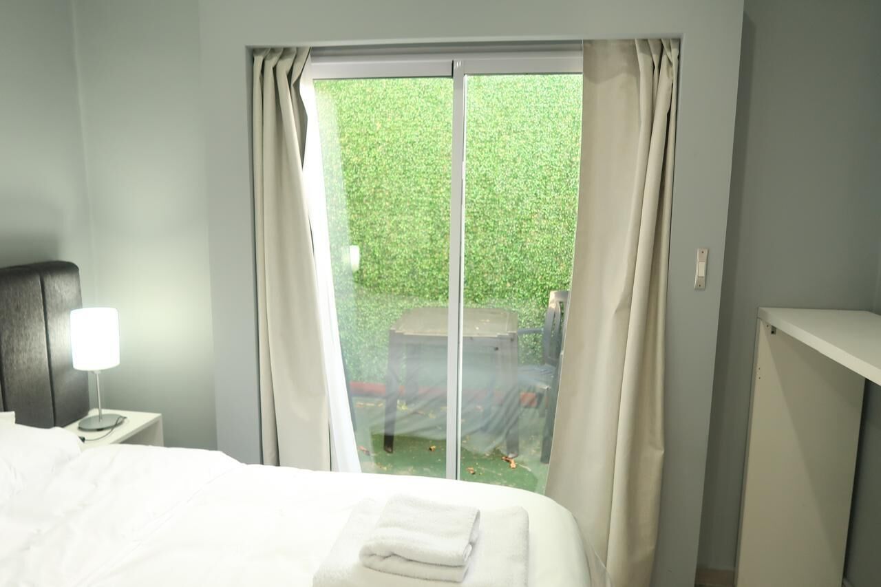 Een hotelkamer met een bed, een nachtkastje met een lamp en uitzicht op een groen buitengebied door een glazen deur.