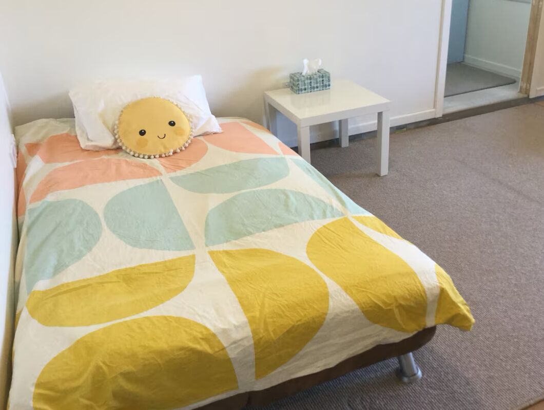 A bed with a colorful bedspread, a small bedside table with a tissue box, and a plain wall.