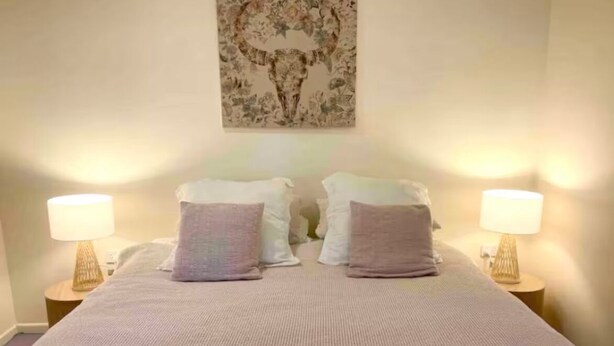 Ein ordentlich bezogenes Bett mit einer rosa Tagesdecke, zwei Kissen und zwei gefalteten Handtüchern. Auf jeder Seite des Bettes stehen zwei Nachttischlampen.