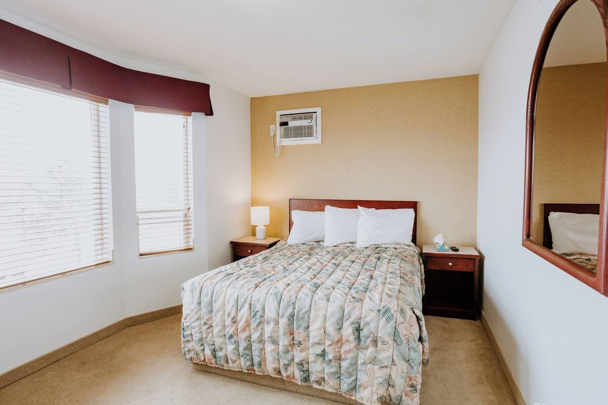 A hotel room with a bed, a nightstand, a lamp, a wall-mounted air conditioner, and a large mirror.
