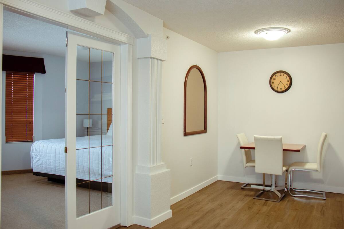 A bedroom with a bed, a door leading to another room, a dining area with a table and chairs, and a wall clock.