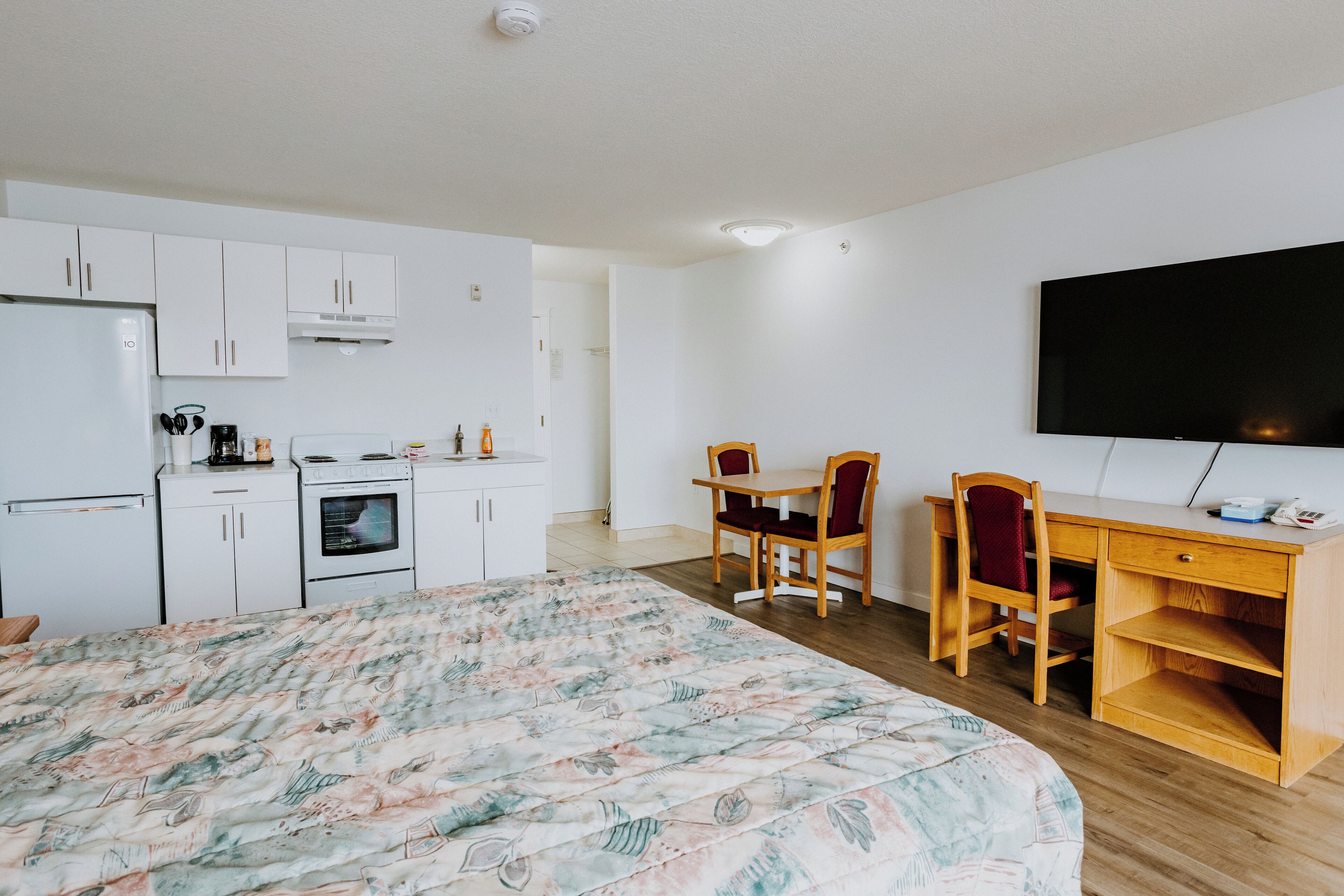 A bedroom with a large bed, a desk, and a TV. There is a kitchen area with cabinets and appliances.