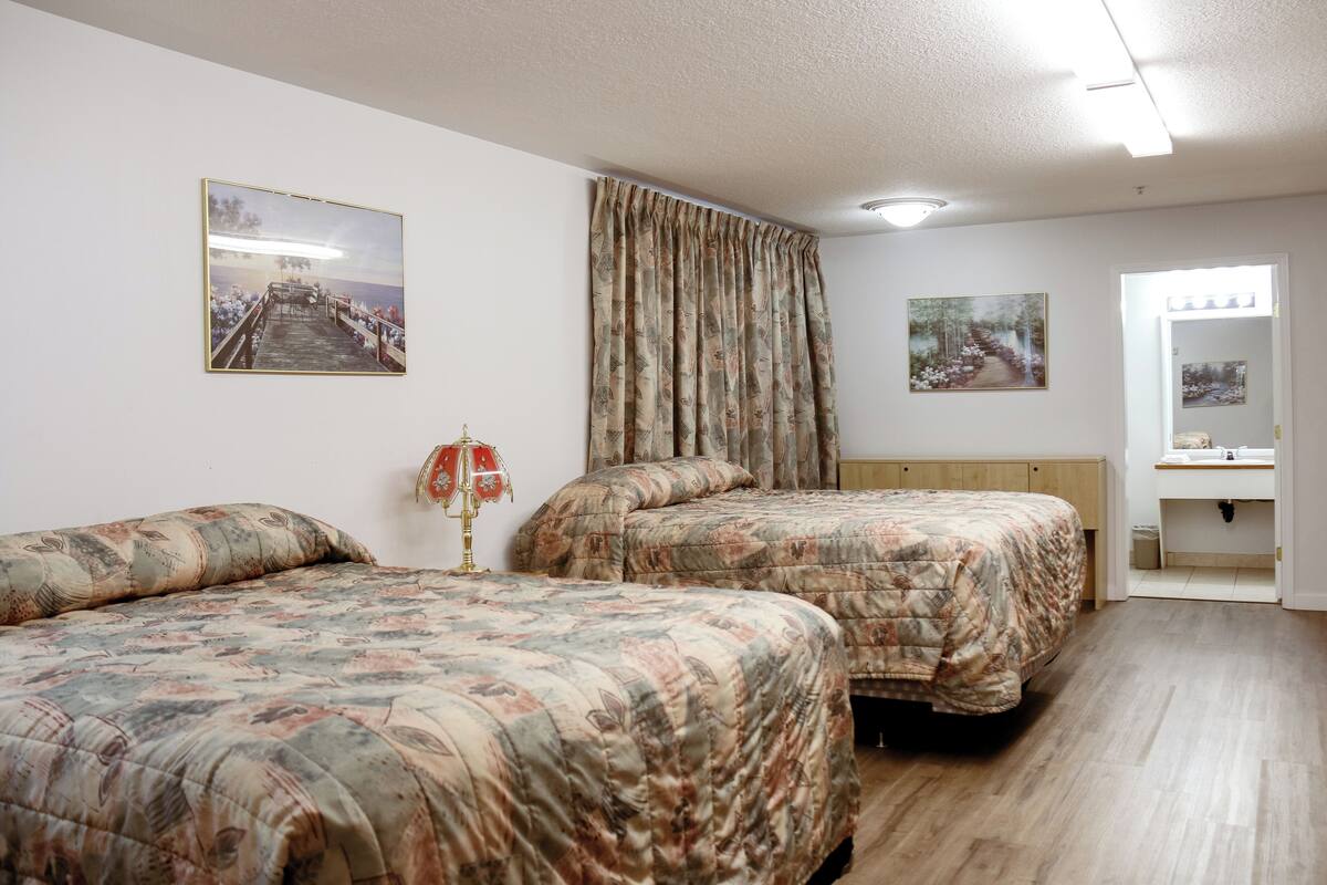 A hotel room with two beds, a wooden floor, a large window with curtains, and a painting on the wall.