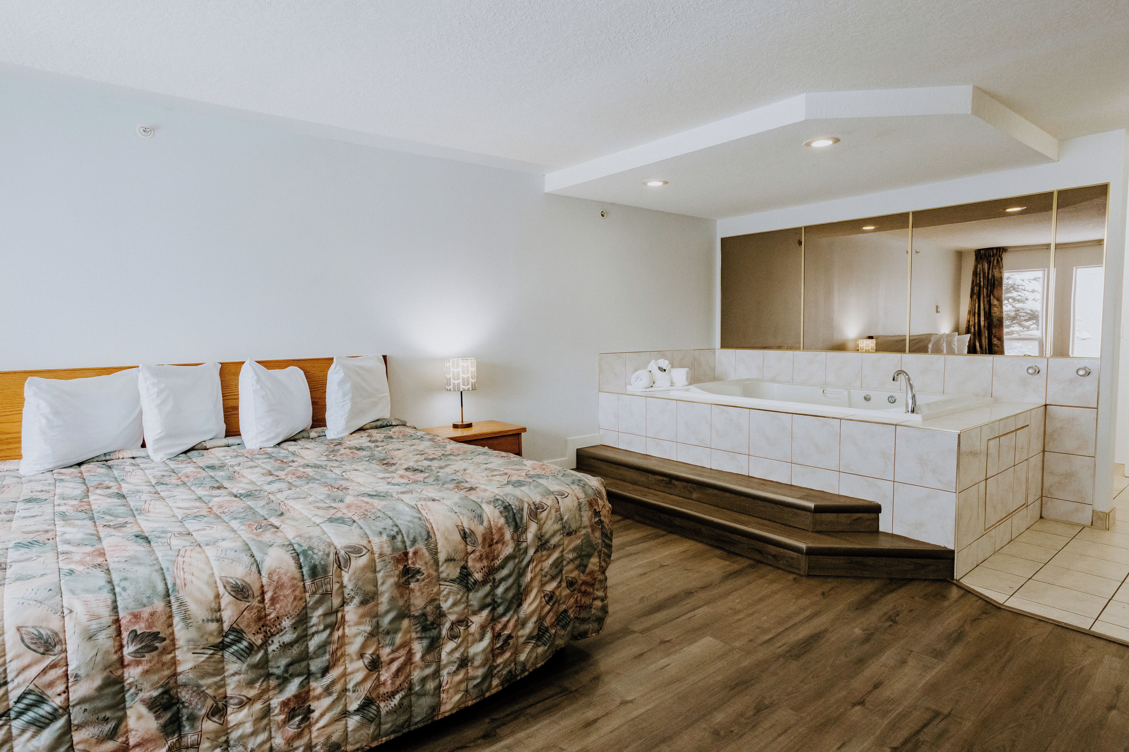 A bedroom with a bed, a nightstand, a large mirror, a built-in bathtub, and wooden flooring.