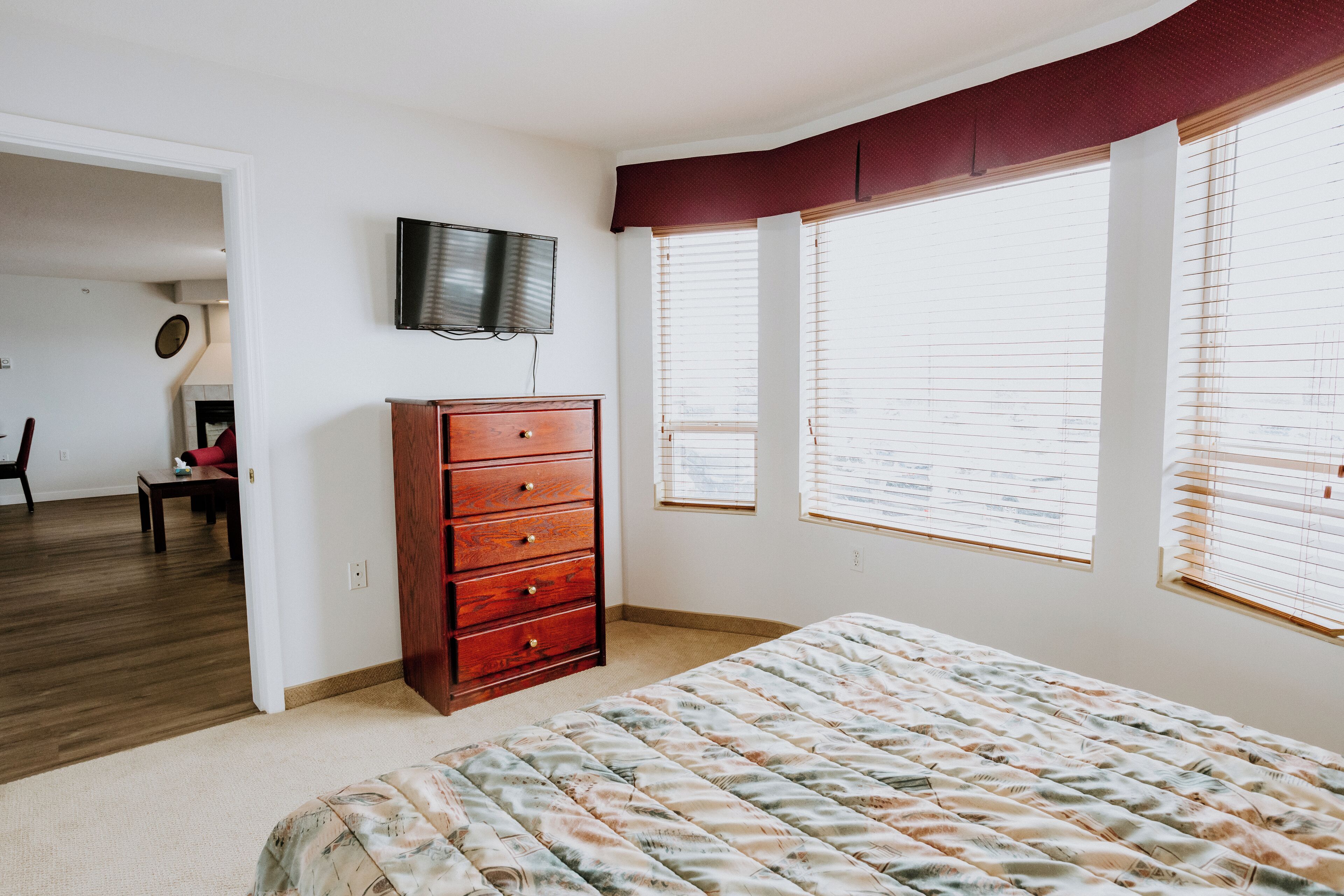 Un dormitorio con cama, cómoda, televisor y dos ventanas con persianas.