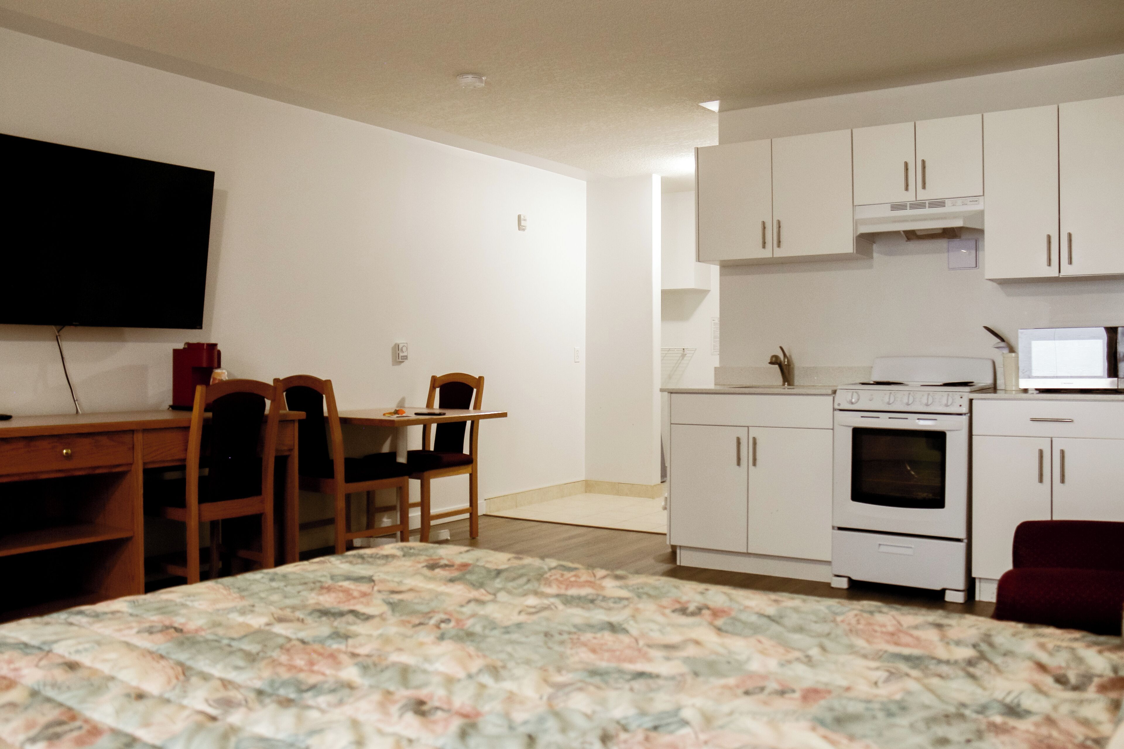 A compact room with a bed, a desk with a chair, a television, a kitchenette with a microwave, and a refrigerator.
