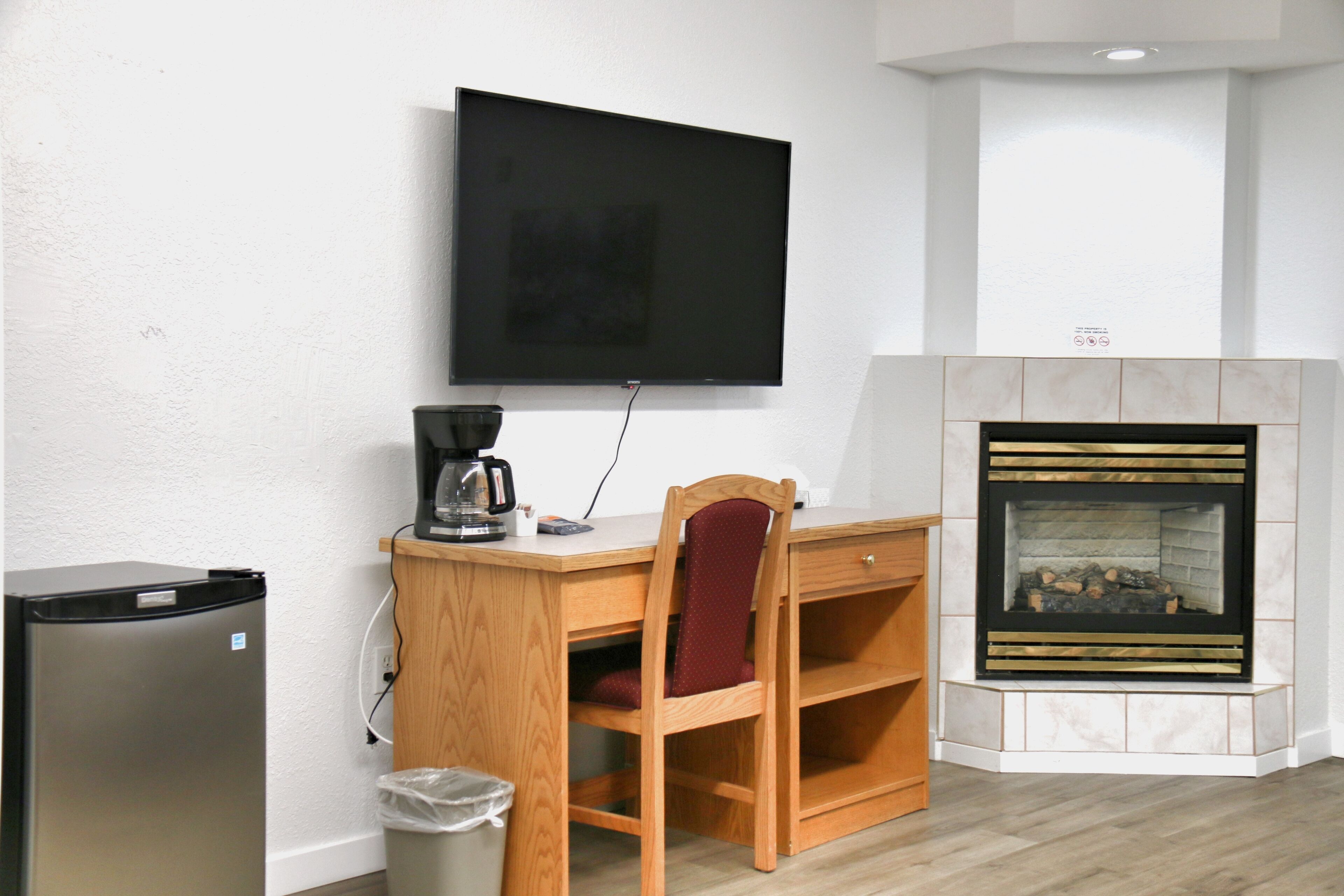 Habitación con televisión de pantalla plana montada en la pared, un escritorio de madera con una silla, cafetera, cesto de basura y chimenea con repisa de piedra.