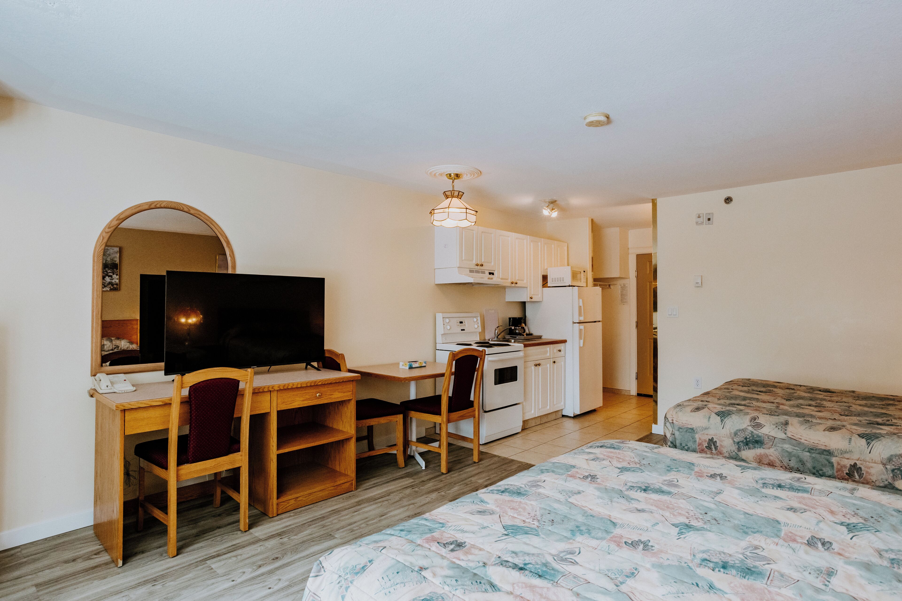 A compact room with a wooden desk, a television, a kitchenette, and a bed with a patterned bedspread.