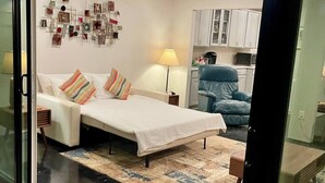 A modern bedroom with a bed, bedside lamps, a blue chair, and a kitchen area in the background. - Hot Tub | 2BR Duplex | Pet Friendly| Near Airport (San Antonio)
