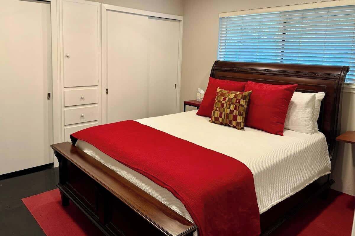 Ein Schlafzimmer mit einem großen Bett, einer hölzernen Bank am Fußende, einem Nachttisch, einer roten und weißen Bettdecke sowie einem Fenster mit Jalousien.