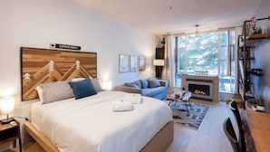 A modern bedroom with a large bed, a sofa, a fireplace, and a view of the outdoors.