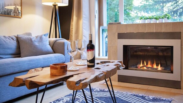 A cozy living room with a sofa, a wooden coffee table, a fireplace, and a large window with a view of trees.