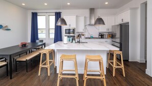 A modern kitchen with white cabinets, a large island with stools, a dining table with benches, and a view of the outdoors through a window.