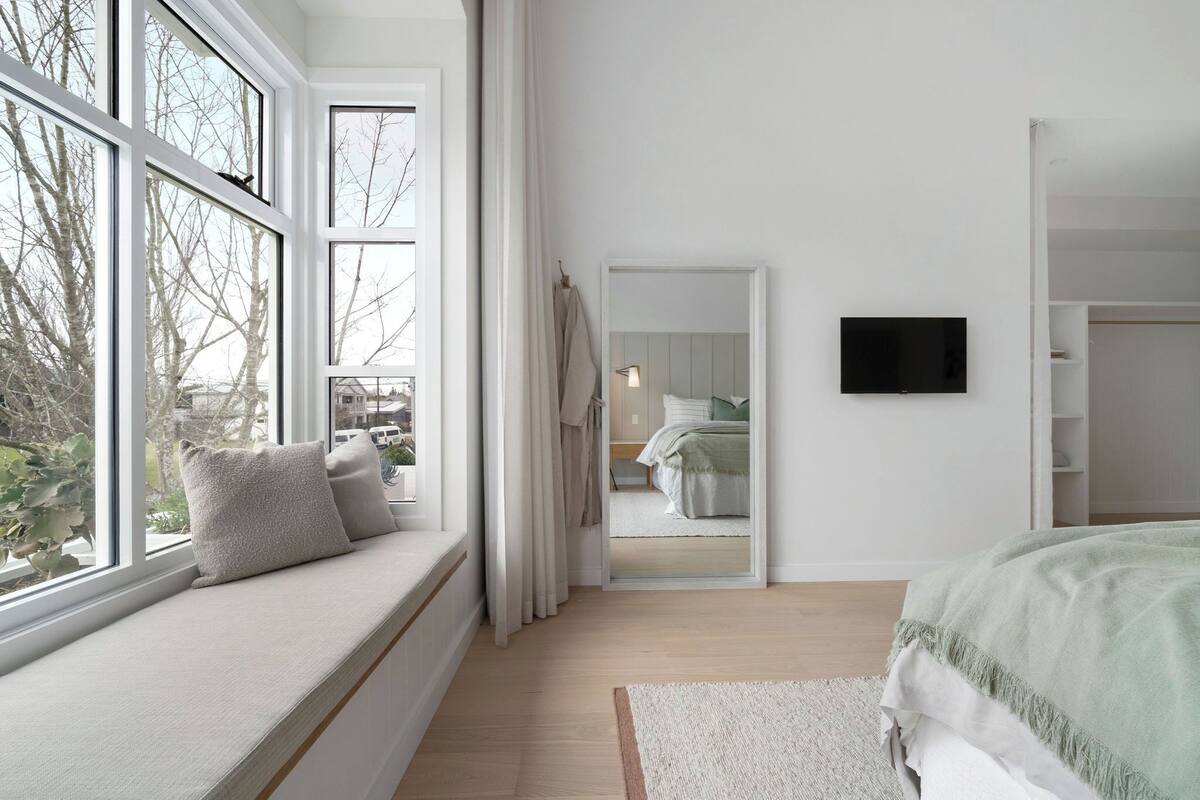A bedroom with a large window, a bed with a green blanket, a bedside table, and a mirror.