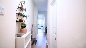 Un pasillo estrecho con una pared blanca, un estante de metal negro con objetos decorativos, una planta en maceta y una puerta blanca al final.