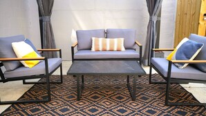 Un espacio exterior moderno con dos sofás grises y una mesa central, rodeado de alfombras estampadas y cortinas.