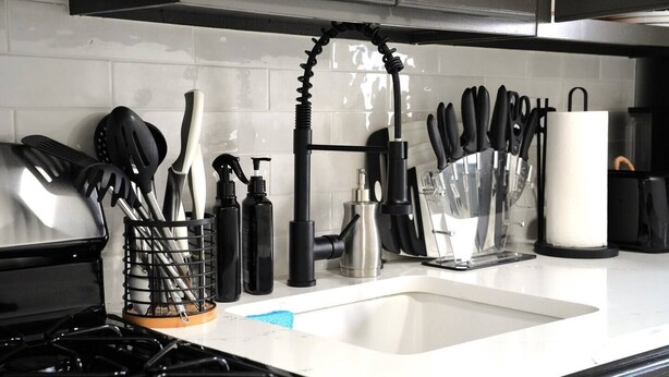 Una cocina con una campana de acero inoxidable, un lavamanos con grifo negro, y diversos utensilios y electrodomésticos de cocina ordenados sobre la encimera.