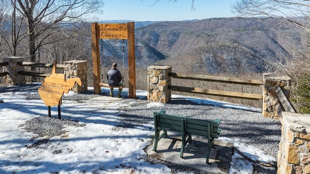 Un panneau en bois portant l’inscription « presque le paradis », placé dans un parc, avec un banc et un pilier en pierre.