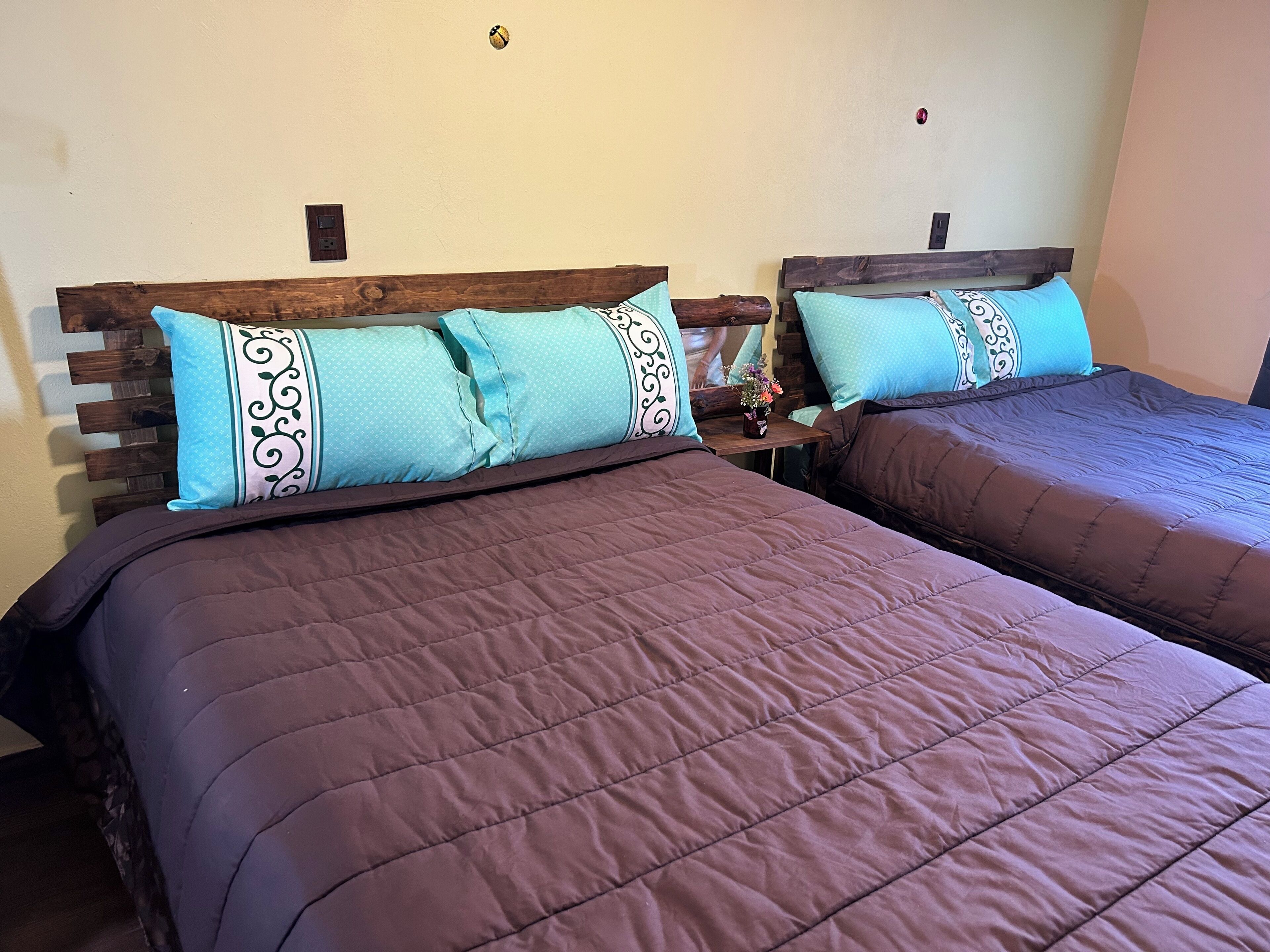 Dos camas con cabeceros de madera y almohadas azules estampadas, una mesita con un florero y un interruptor de luz en la pared.
