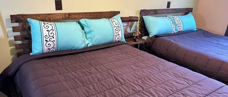 Dos camas con cabeceros de madera y almohadas azules estampadas, una mesita con un florero y un interruptor de luz en la pared.