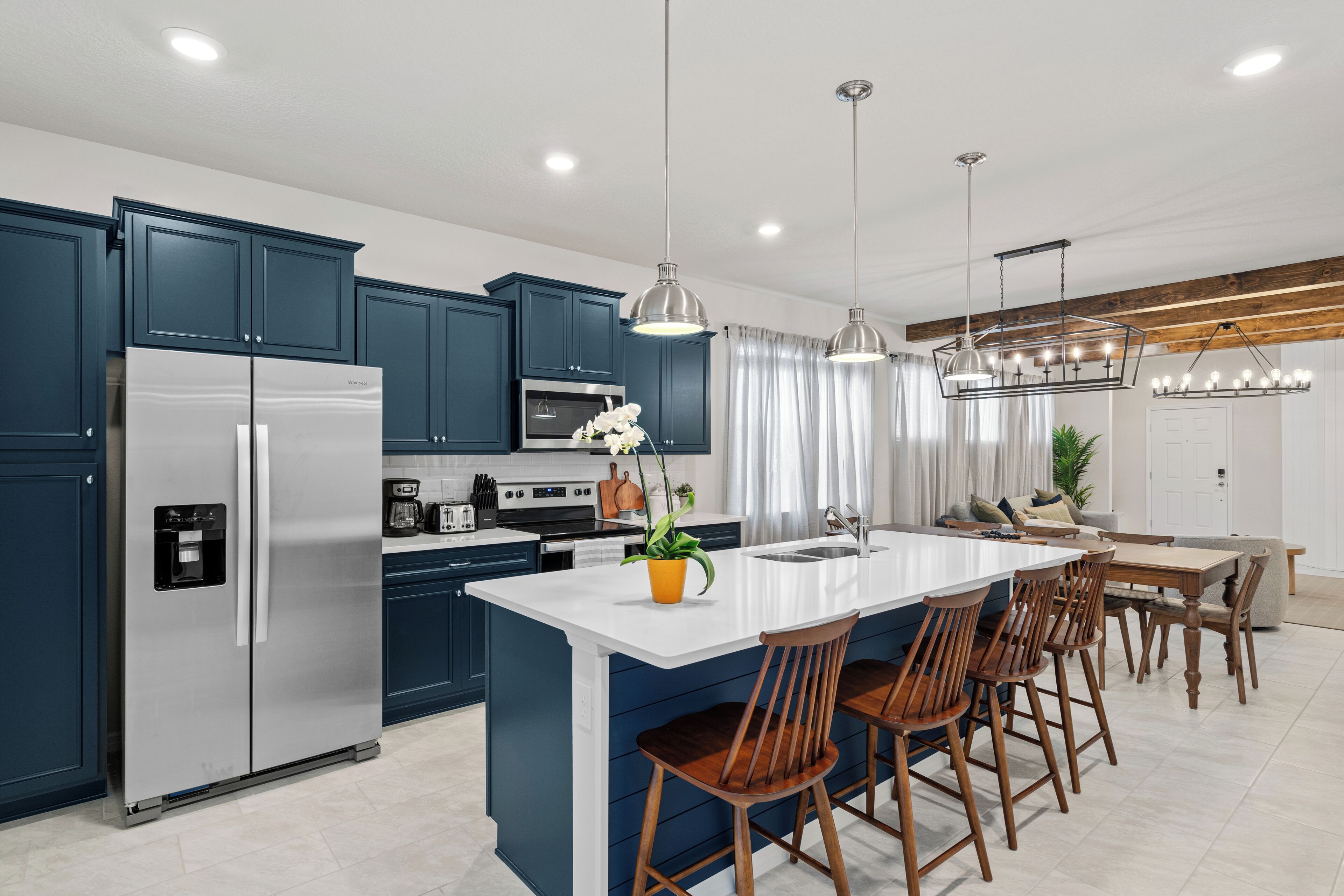 A modern kitchen with dark blue cabinets, stainless steel appliances, a central island with bar stools, pendant lighting, and a dining area with a wooden table and chairs.